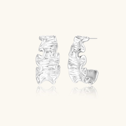 White gold plated sculptural hoop earrings with organic wavy edges and reflective metallic finish on cream background